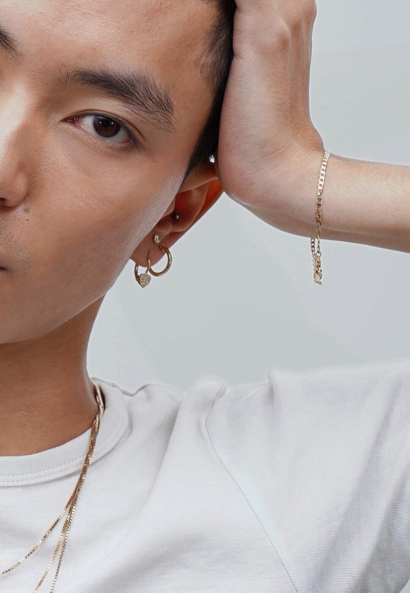 Gold jewelry features include hoop earrings, a heart-shaped charm, and a slender chain bracelet. The wearer has short hair and a plain white shirt.