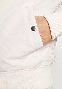 Ľahká krémová bomber bunda s rebrovanými manžetami, ktorá má strieborný cvok na rukáve a je vyrobená z hladkého syntetického materiálu.