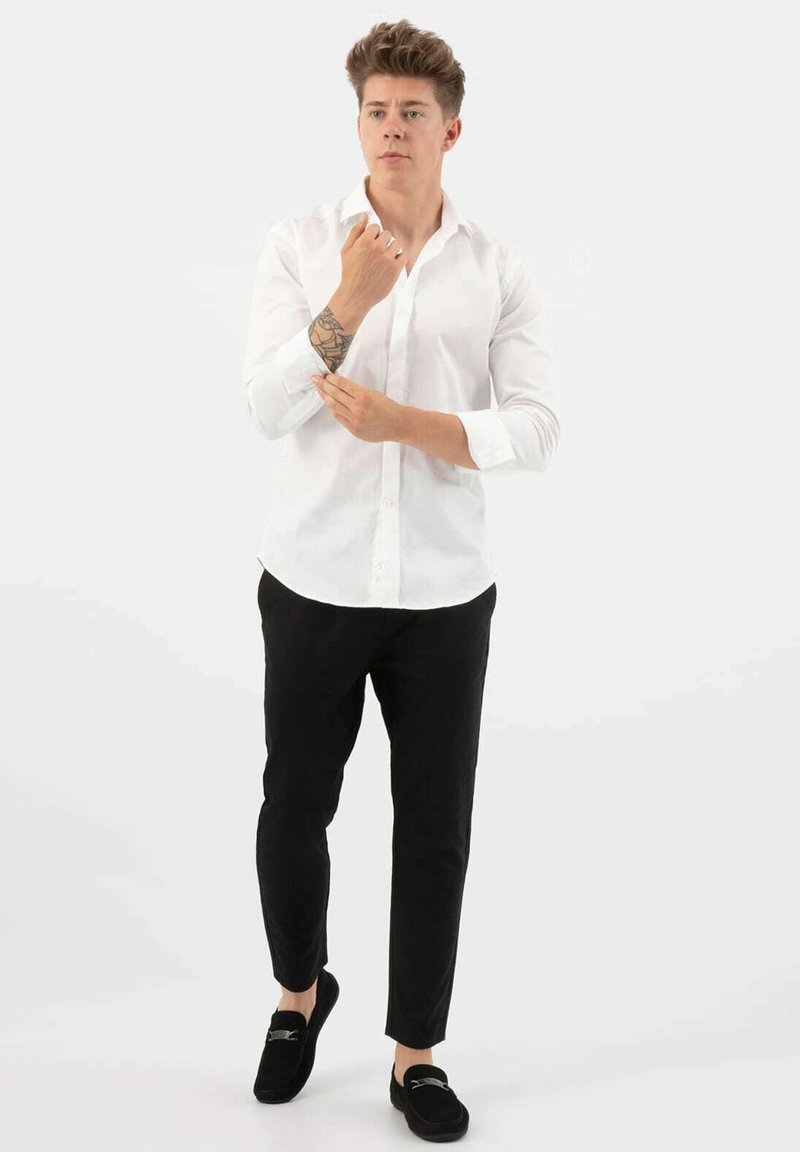 Witte overhemd met knopen, zwarte slim-fit broek en zwarte loafers. Model met een tatoeage, met opgestoken mouwen van het shirt, staand tegen een effen achtergrond.