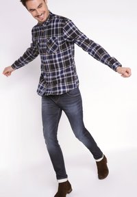 Ruudullinen tummansininen ja musta flanellipaita, jossa on napitus edessä, kaksi rintataskua, yhdistettynä tummiin farkkuihin ja ruskeisiin mokkasiinivaljaisiin.