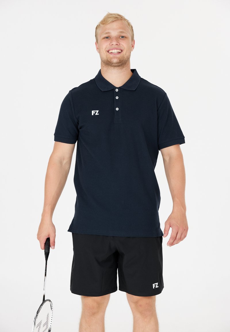 Marinblå pikétröja med krage, tre knappar och vit "FZ"-logotyp. Matchas med svarta shorts. Modellen håller i ett badmintonracket.