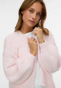 Svijetlo ružičasti pleteni kardigan s labavim, teksturiranim materijalom i rebrastim manžetama. Model drži kardigan, prikazujući mekan, oversized dizajn.