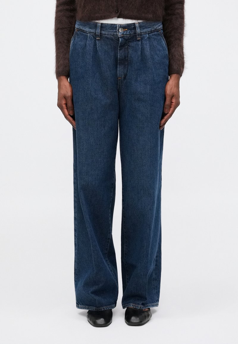 Jeans de mezclilla azul de pierna ancha con cintura alta, que presentan pliegues frontales y un diseño clásico de cinco bolsillos. Textura suave y de peso medio.