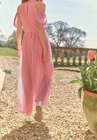 Robe maxi en mousseline rose avec manches épaules dénudées et design à couches, portée avec des chaussures blanches, sur fond de jardin.