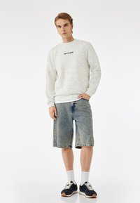 Sudadera texturizada gris claro con logo negro, combinada con pantalones cortos de mezclilla desgastados hasta la rodilla. Terminado con zapatillas negras y calcetines blancos.