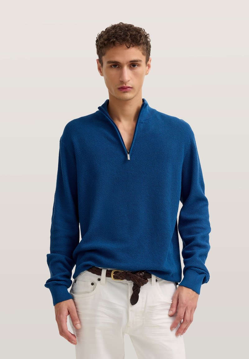 Young man wearing a blue quarter-zip sweater and white pants with a brown braided belt, standing against a neutral background.