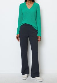 Pull en tricot côtelé vert avec un col en V, associé à un pantalon évasé gris foncé et des baskets blanches. Texture lisse et coupe décontractée.