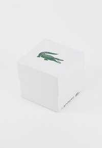Vit presentask med texturerad yta, med en grön Lacoste krokodil-logotyp på toppen och varumärkesnamn på sidan. Fyrkantig form.