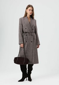 Lange, dubbelgeknoopte jas in bruin pied-de-pouleweefsel, met riem in de taille. Gecombineerd met een donker suède handtas en zwarte kniehoge laarzen.