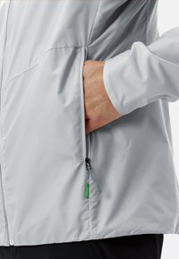 Veste gris clair avec une poche zippée. Tissu lisse et léger avec un subtil éclat. Comprend une étiquette verte et un cordon de serrage ajustable.