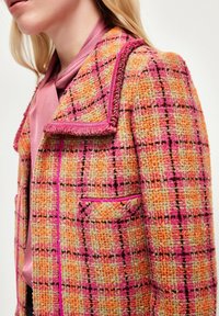 Veste en tweed multicolore texturée présentant un motif à carreaux orange, jaune et rose, avec un col à franges et des détails d'accentuation roses.