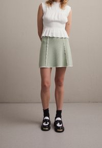 Weißes, gefaltetes Oberteil mit hohem Kragen, kombiniert mit einem mintgrünen A-Linien-Rock mit gewellten Kanten und dekorativen Nahtakzenten. Schwarze Schuhe.