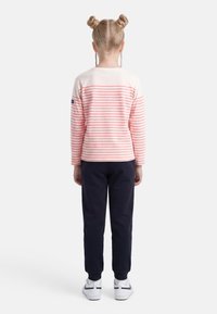 Παιδί με ξανθά μαλλιά δεμένα σε διπλούς κότσους, φοράει ροζ και λευκή ριγέ μπλούζα με μακριά μανίκια, σκούρο μπλε παντελόνι και άσπρα αθλητικά παπούτσια, στέκεται με την πλάτη προς τα εμπρός.