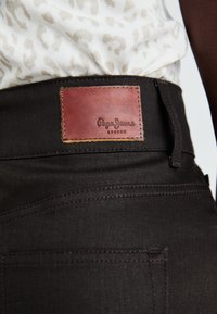 Jeans de culoare maro închis, dintr-un material texturat, având o etichetă din piele inscripționată "Pepe Jeans London" și detalii de cusătură vizibile.