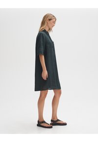 Diep groen linnen jurk met korte mouwen, een kraag en een borstzak, die net boven de knieën valt. Gecombineerd met zwarte platte sandalen.