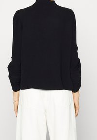 Top corto nero a maniche lunghe con spalle a palloncino, caratterizzato da un tessuto testurizzato e dettagli di cucitura discreti. Pantaloni bianchi a vita alta visibili sotto.