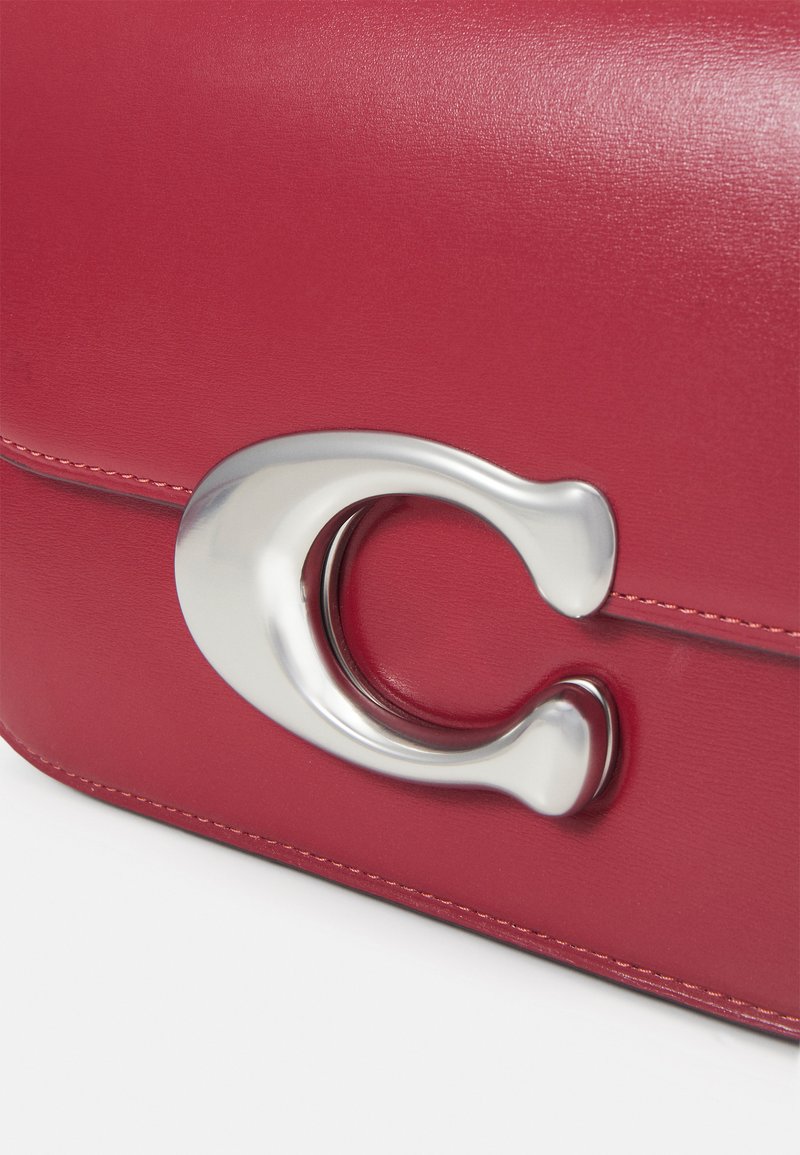Rote Lederhandtasche mit glatter Textur, ausgestattet mit einem auffälligen silbernen Hardwareverschluss im kreisförmigen Design und kontrastierenden Nähten.