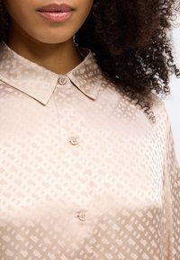 Ανοιχτό ροζ μπ�λουζάκι με σχέδιο και γιακά, με γεωμετρική υφή και δύο κουμπιά μπροστά. Λείος ύφασμα με διακριτική γυαλάδα.