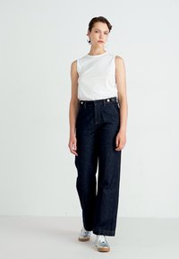 Top bianco senza maniche abbinato a pantaloni a gamba larga in denim scuro a vita alta. Gli accessori includono grandi orecchini a cerchio e sneakers argentati in stile retrò.