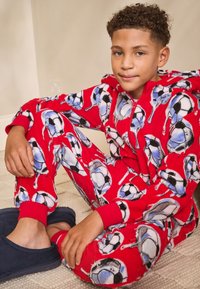 Tuta intera in pile rosso con motivo a pallone da calcio, con chiusura a zip e polsini a costine rossi. Indossata con pantofole blu.