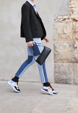Blazer noir à fines rayures porté sur une chemise bleu clair, associé à un jean en denim patchwork avec bords effilochés, et des baskets noires et blanches.