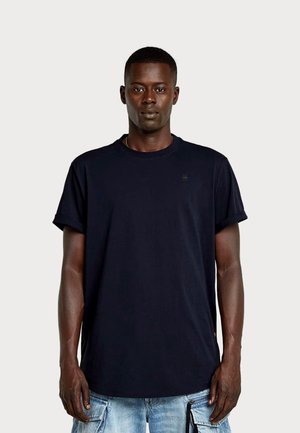Ung mand med mørk hud og kort hår iført en ensfarvet sort t-shirt og lyseblå cargoshorts, stående mod en ensartet baggrund.
