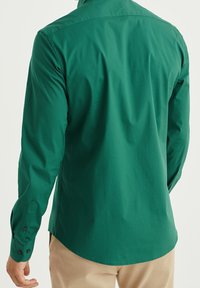 Grön långärmad skjorta i en solid färg, tillverkad av ett slätt tyg, med knäppta manschetter och en rundad nederkant vid midjan.