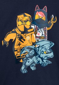 Dva LEGO ninja lika, jedan u narančastom oklopu, a drugi u bijelom s mačjim ušima, stoje iza malog plavog zmaja s krilima.