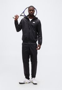 Άνδρας με μαύρα αθλητικά ρούχα κρατώντας ρακέτα τένις πάνω στον ώμο, φοράει λευκά παπούτσια EA7, στέκεται μπροστά σε απλό φωτεινό φόντο.