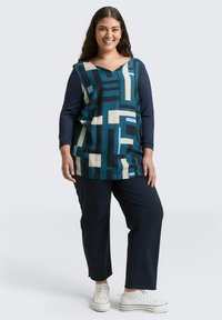 Langsleeve top met een geometrisch patroon in teal, crème en marineblauw, gecombineerd met marineblauwe broek en witte sneakers. Zacht, vallend materiaal.