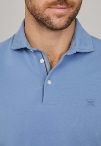 Gaiši zilā polo krekls, kas izgatavots no teksturētas auduma, ar divu pogu apvalku un izšūtu logo uz kreisās krūtis.