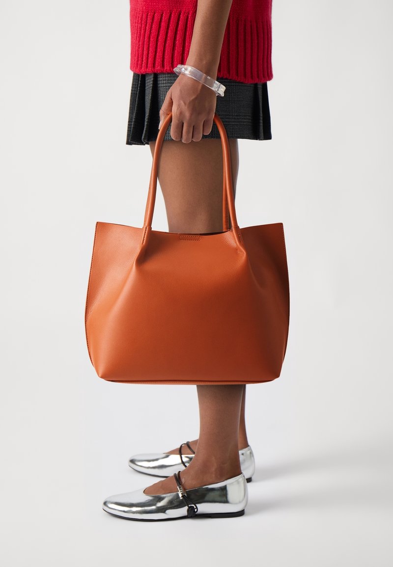 Personne tenant un grand sac fourre-tout en cuir marron, portant un pull rouge, une jupe plissée noire, un bracelet transparent et des chaussures Mary Jane argentées, debout sur fond blanc.