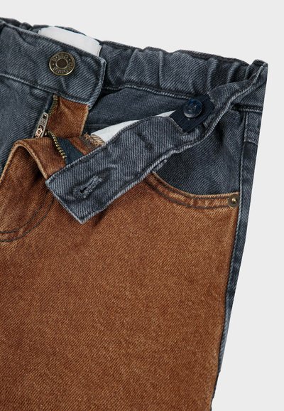 Traperice s tamnoplavim pojasom i svijetlosmešnim prednjim džepom. Zipper i dugme su vidljivi, s kontrastnim teksturama.