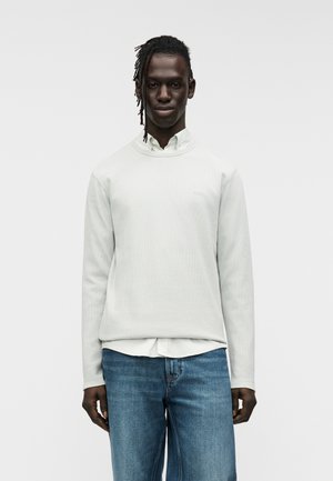 Γκρι ανοιχτόφωμο φουτεράκι με υφή και στρογγυλή λαιμόκοψη, με μικρό λογότυπο της μάρκας. Φορεμένο πάνω από ένα λευκό πουκάμισο με γιακά και συνδυασμένο με μπλε τζιν.