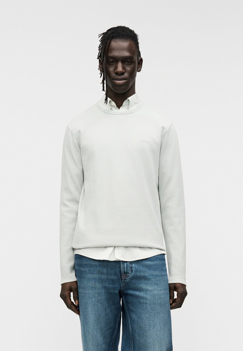 Γκρι ανοιχτόφωμο φουτεράκι με υφή και στρογγυλή λαιμόκοψη, με μικρό λογότυπο της μάρκας. Φορεμένο πάνω από ένα λευκό πουκάμισο με γιακά και συνδυασμένο με μπλε τζιν.