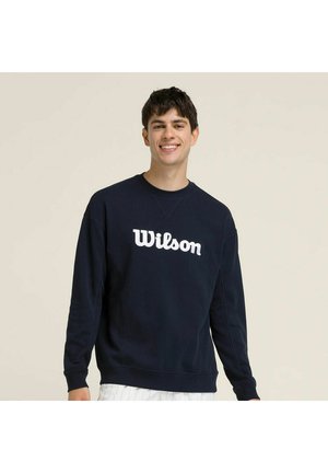 Giovane uomo sorridente, indossa una felpa nera Wilson e pantaloni bianchi su uno sfondo chiaro e uniforme.