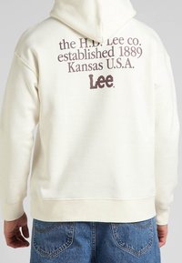 Sudadera de color crema con un gran estampado gráfico marrón en la parte posterior que incluye texto y un logotipo. La tela tiene una apariencia suave y texturizada.