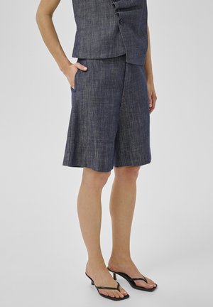 Persoon draagt donkerblauwe denim short tot op de knie, bijpassende mouwloze top met knopen en zwarte sandalen met hak en teenslipper, staand met hand in de zak.