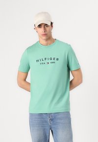 Mintgrønn t-skjorte med korte ermer, med et navy-logo og årstall trykt på. Kombinert med lyseblå jeans og en beige caps.