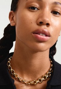 Collana in oro con maglie lisce e intrecciate. Finitura lucida con forme arrotondate, indossata sopra una maglia nera con colletto.