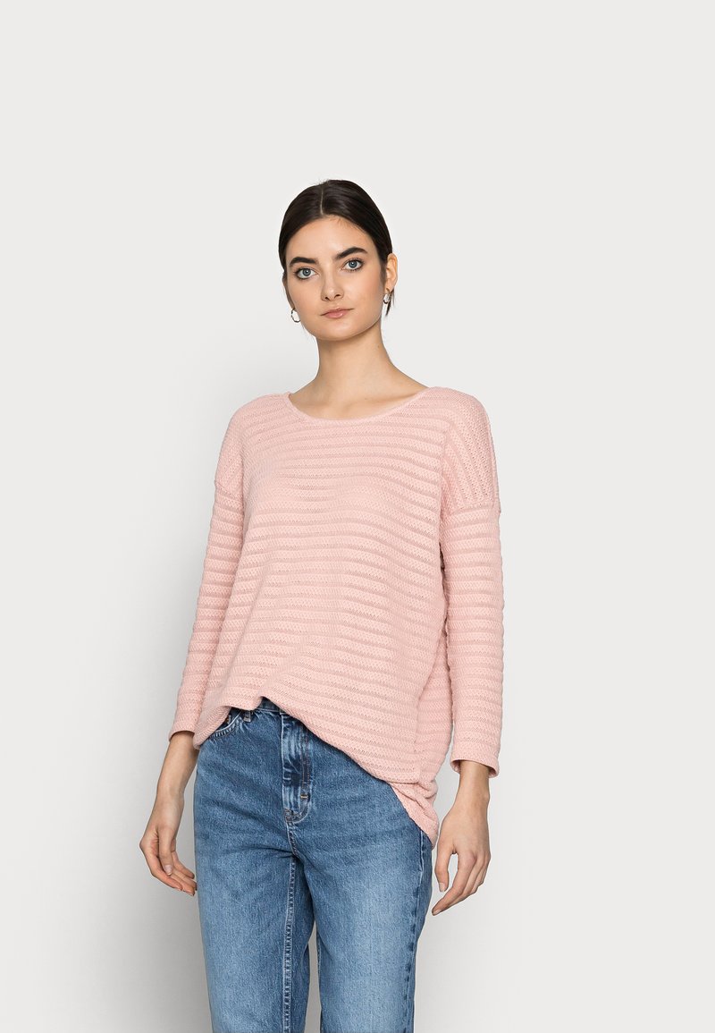 Sweater de malha rosa claro com um padrão riscado texturizado, decote largo e barra traseira ligeiramente mais longa, combinado com jeans azul claro.