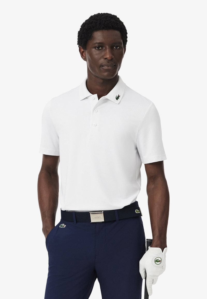 Hombre con camisa polo blanca, pantalones azul marino, cinturón a juego y guante de golf blanco, sosteniendo un palo de golf, de pie frente a un fondo blanco.