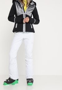 Schwarz-graue Skijacke mit Kapuze aus künstlichem Fell, Reißverschlusstaschen und kontrastierendem Material. Kombiniert mit weißen Hosen und skischuhen mit grünen Akzenten.