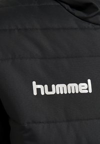 Sort jakke med quiltet tekstur, der har et fremtrædende hvidt "hummel" logo. Stoffet ser glat ud med en let glans.