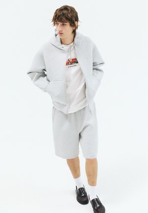 Junger Mann trägt einen hellgrauen Hoodie, passende Shorts, weißes T-Shirt mit Grafik, weiße Socken und schwarze Loafers, steht vor weißem Hintergrund.