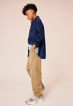 Denim-Hemd mit Knopfleiste und zwei Brusttaschen, khakifarbene Hose und graue Sneakers mit weißen Akzenten. Das Modell steht mit den Händen in den Taschen.