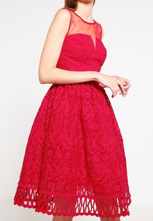 Robe rouge sans manches en dentelle avec motif floral, encolure transparente, taille cintrée et détail échancré à l'ourlet portée par une femme debout les mains claspées.