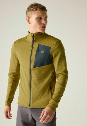 Mann trägt olivgrüne Jacke mit dunkler Brusttasche, steht und schaut seitlich vor einem schlichten hellen Hintergrund.