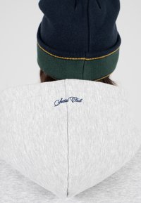 Felpa grigia con cappuccio e berretto navy blu e verde. La felpa ha la dicitura "Special Club" ricamata in blu navy sul collo posteriore.