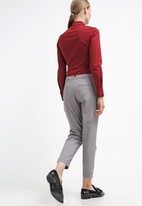 Chemise rouge ajustée avec col pointu, associée à un pantalon gris clair sur mesure et des chaussures noires. Tissu lisse, design élégant et longueur raccourcie.